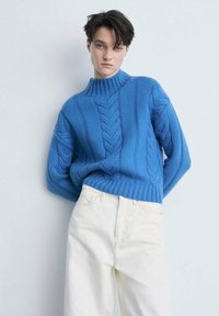 Blauer Strickpullover mit hohem Kragen und Zopfmuster. Kombiniert mit hellen, hochgeschnittenen Hosen. Schlichtes, strukturiertes Design.