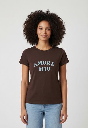 Giovane donna con capelli ricci che indossa una maglietta marrone con la scritta "AMORE MIO" e jeans azzurri, in piedi davanti a uno sfondo semplice.