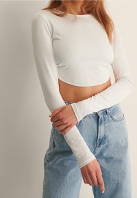 Weißes langärmliges Cropped-Top aus glattem Stoff mit rundem Halsausschnitt. Kombiniert mit hoch taillierten hellblauen Jeans.