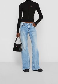 Henkilö, joka on pukeutunut mustaan vetoketjulliseen huppariin ja vaaleansiniisiin leveälahkeisiin farkkuihin, pitää kädessään mustaa käsilaukkua, jossa on logo-nauha.