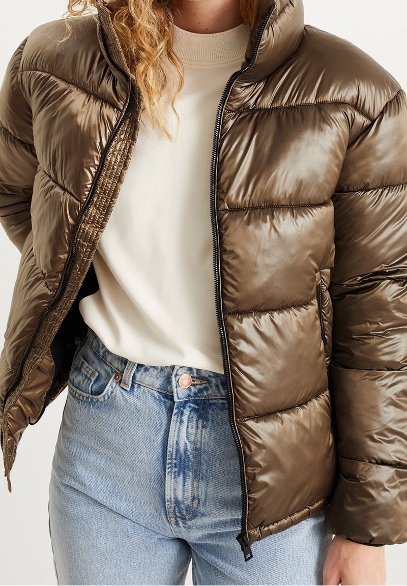 Person med en skinnende brun dunjakke, der er åbnet, over en cremefarvet sweater og lyseblå højtaljede jeans, står foran en hvid baggrund.
