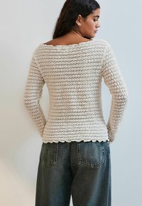 Kremfarvet strikket sweater med bred halsudskæring, lange ærmer og bølgekant, som er parret med højtaljede, løstsiddende grå jeans.