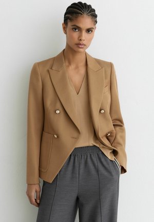 Tan dobbeltradet blazer med tekstureret stof, hakkede revers, knapper i guldfinish, kombineret med en beige top og grå bukser med snøre.