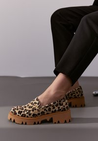 Schwarze Slip-On-Schuhe mit Leopardenmuster und einer dicken, strukturierten braunen Sohle mit gezacktem Rand sowie einem glatten Obermaterial.