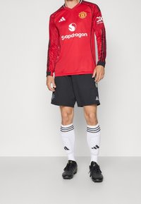Maglia rossa a maniche lunghe con dettagli neri, presenta un logo e il nome dello sponsor. Pantaloni neri e calze bianche con tre strisce nere, abbinati a scarpini neri.