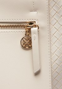 Section d'un sac en cuir crème avec une fermeture éclair métallique, des accents dorés et un charme décoratif. Présente une texture tressée en relief sur le côté.