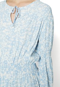 Vestido azul com padrão floral, apresentando um decote em forma de olho de cerradura, cintura elástica e mangas longas plissadas, confeccionado em tecido leve com uma textura suave.
