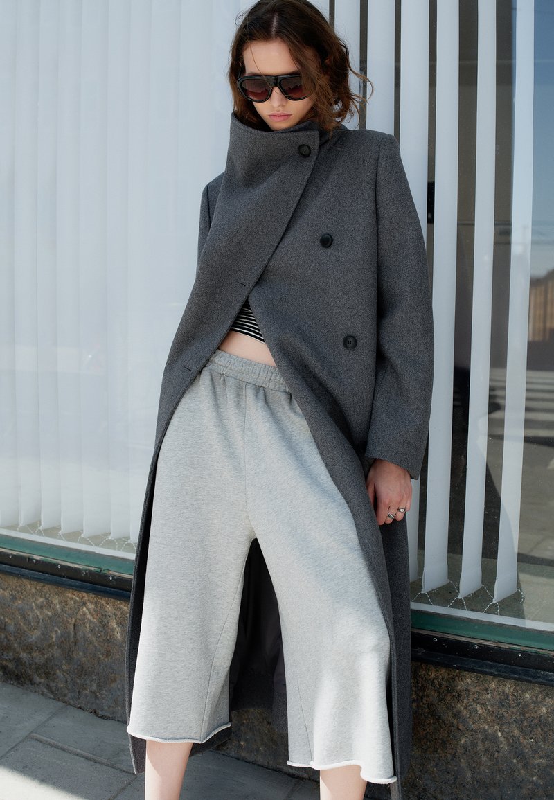 Femme portant un manteau gris oversize, un pantalon gris à jambes larges et des lunettes de soleil, debout contre une fenêtre en verre avec des stores verticaux.