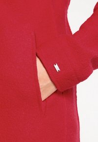 Rode wollen jas met een gestructureerde afwerking, voorzien van een zijzak en een zilveren merklabel nabij de manchette.