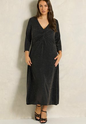 Femme portant une longue robe noire étincelante avec des manches trois-quarts et un décolleté en V, assortie à des sandales à talons hauts noires à brides.