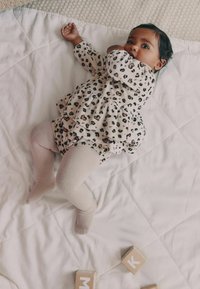 Leopard print dress in beige and black, light beige tights, and hand raised; child lying on a white quilt with wooden blocks nearby.