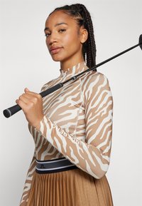 Dugi rukav golf majica u bež boji s bijelim prugama zebre, s visokom ovratnikom i kontrastnim šavovima. Model drži golf palicu.