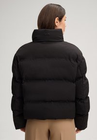 Giacca a vento nera, design trapuntato con colletto alto, caratterizzata da un sottile dettaglio del logo sul retro. Tessuto liscio e vestibilità oversize.