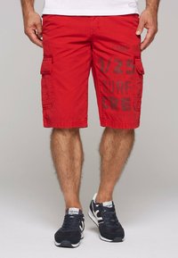 Rode cargoshorts met zijzakken, voorzien van een zwart gedrukte tekstontwerp. Gdragen met marineblauwe sneakers en een wit T-shirt. Gladde katoenmateriaal.