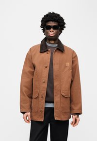 Καφέ βαμβακερό μπουφάν με γιακά από κοτλέ, που διαθέτει δύο μεγάλες μπροστινές τσέπες και κουμπιά με τσαμπί, φορεμένο πάνω από γκρι φούτερ και μαύρο παντελόνι.