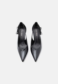 Chaussures à talons hauts en cuir verni noir avec un bout pointu et un design avec découpe. Elles présentent de petits accents de nœuds à l'arrière et une texture lisse.