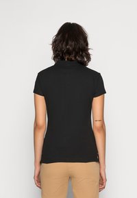 Persona con capelli mossi scuri, che indossa una polo nera a maniche corte e pantaloni beige, vista da dietro su uno sfondo semplice.