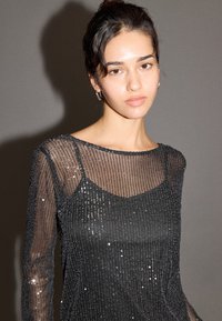 Svart långärmad topp i genomskinligt tyg med paljettprydda vertikala ränder, med rund halsringning och smal passform, som visar en slät textur.