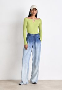 Top verde a costine senza spalle con dettagli a chiave, abbinato a jeans in denim larghi di un blu chiaro. La modella indossa un cappello beige e scarpe nere.