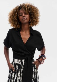 Zwarte blouse met een col, gecombineerd met hoog-waist zwarte en witte gebloemde broeken. Accenten omvatten kleurrijke armbanden om de pols.