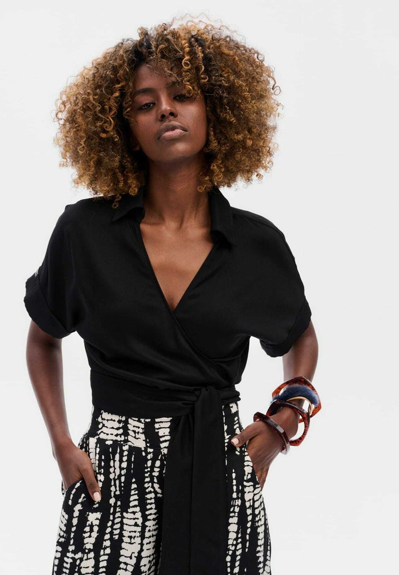 Zwarte blouse met een col, gecombineerd met hoog-waist zwarte en witte gebloemde broeken. Accenten omvatten kleurrijke armbanden om de pols.