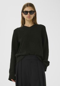 Black, textured v-neck sweater with long sleeves, paired with high-waisted, dark trousers. Model wearing angular black sunglasses.