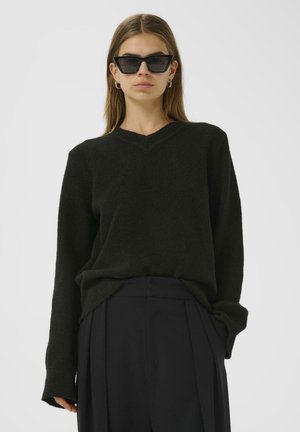 Black, textured v-neck sweater with long sleeves, paired with high-waisted, dark trousers. Model wearing angular black sunglasses.