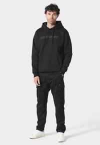 Svart hettegenser med hette, med en subtil logo og frontlomme. Parret med svarte cargo-bukser og hvite sneakers. Flatt, uformelt design.