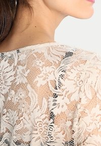 Top in pizzo con motivo floreale, colore crema, caratterizzato da un design in rete aperta e dettagli intricati. Texture morbida e scollatura rotonda.