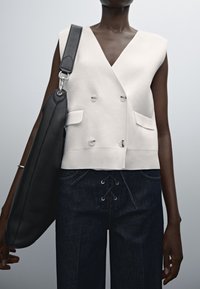 Osoba o ciemnej karnacji, ubrana w bezrękawnik w kolorze białym, dwurzędowy, ciemne spodnie sznurowane, niosąca dużą czarną skórzaną torbę na ramię.