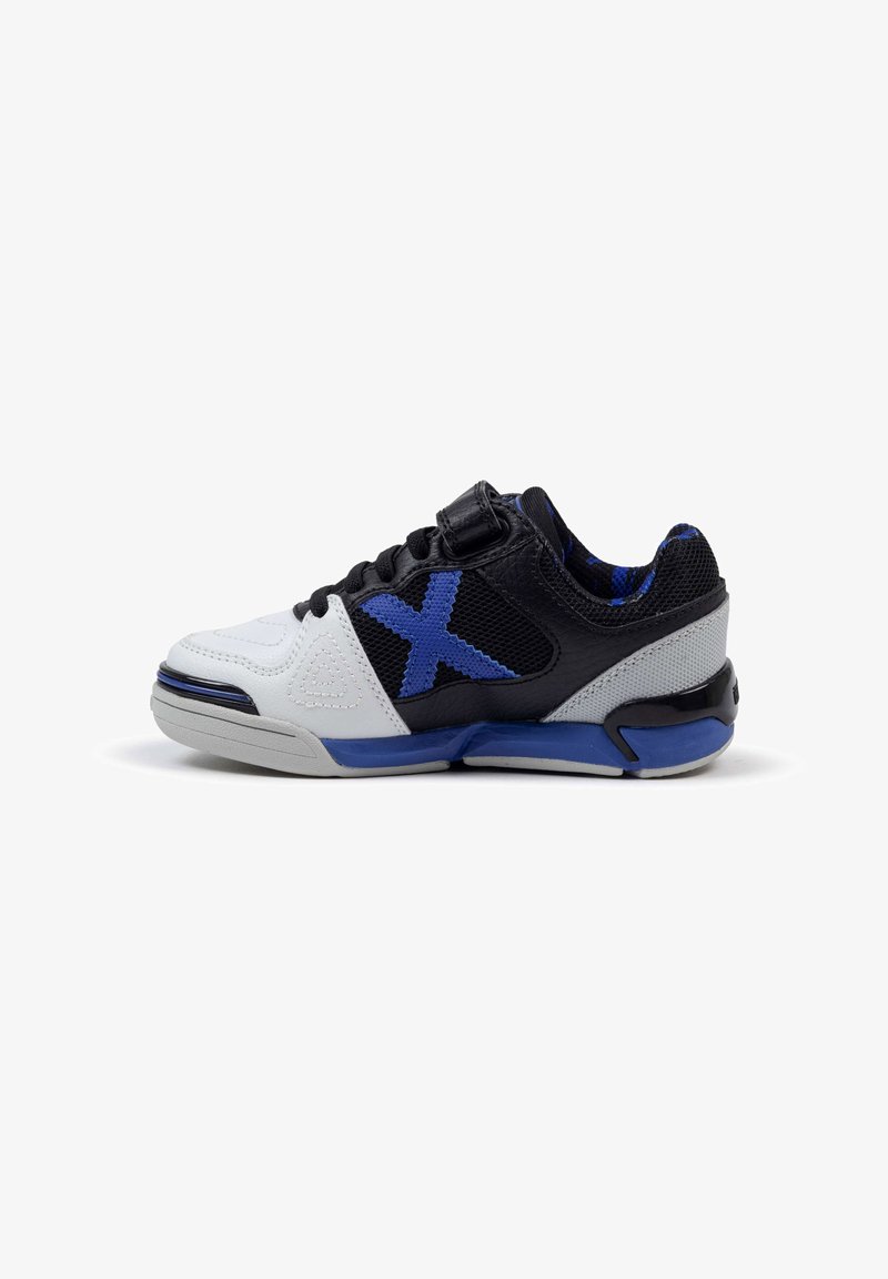 Zapatilla deportiva en blanco y negro con acentos azules, hecha de materiales de malla y cuero, con puntera redondeada, correa de velcro y superficie texturizada en los laterales.