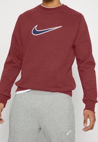 Bordeauxfärgad sweatshirt med en stor vit och marinblå Nike swoosh-logotyp. Har rundad halsringning och muddar. Kombineras med gråa mjukisbyxor.