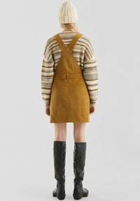 Geel corduroy-jurkje met gekruiste banden, gecombineerd met een multicolor gestreept sweater en kniehoge zwarte leren laarzen.