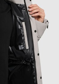 Giacca isolante grigio chiaro con interno nero lucido. Presenta bottoni a pressione, zip e un'etichetta interna con testo bianco.