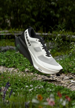 Chaussure de course Mammut blanche et noire suspendue au-dessus d'un sentier extérieur rocheux et herbeux avec un feuillage vert en arrière-plan.