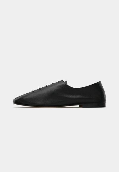 Chaussure en cuir noir à bout pointu, design minimaliste, ornée de petits clous décoratifs le long de la tige. Semelle plate sans motifs visibles.