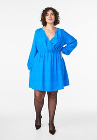 Zilas ietīšanas kleita ar garām piedurknēm, strukturētu audumu un iespaidīgu vidukļa sašaurinājumu. Atšķirīgās iezīmes ir dziļš V veida kakla izgriezums un izplestos svārki.