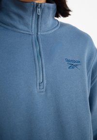 Ljusblå fleecetröja med halv dragkedja och hög krage, med texturerad yta och broderad logotyp på vänster bröst. Dragkedjan matchar färgen.