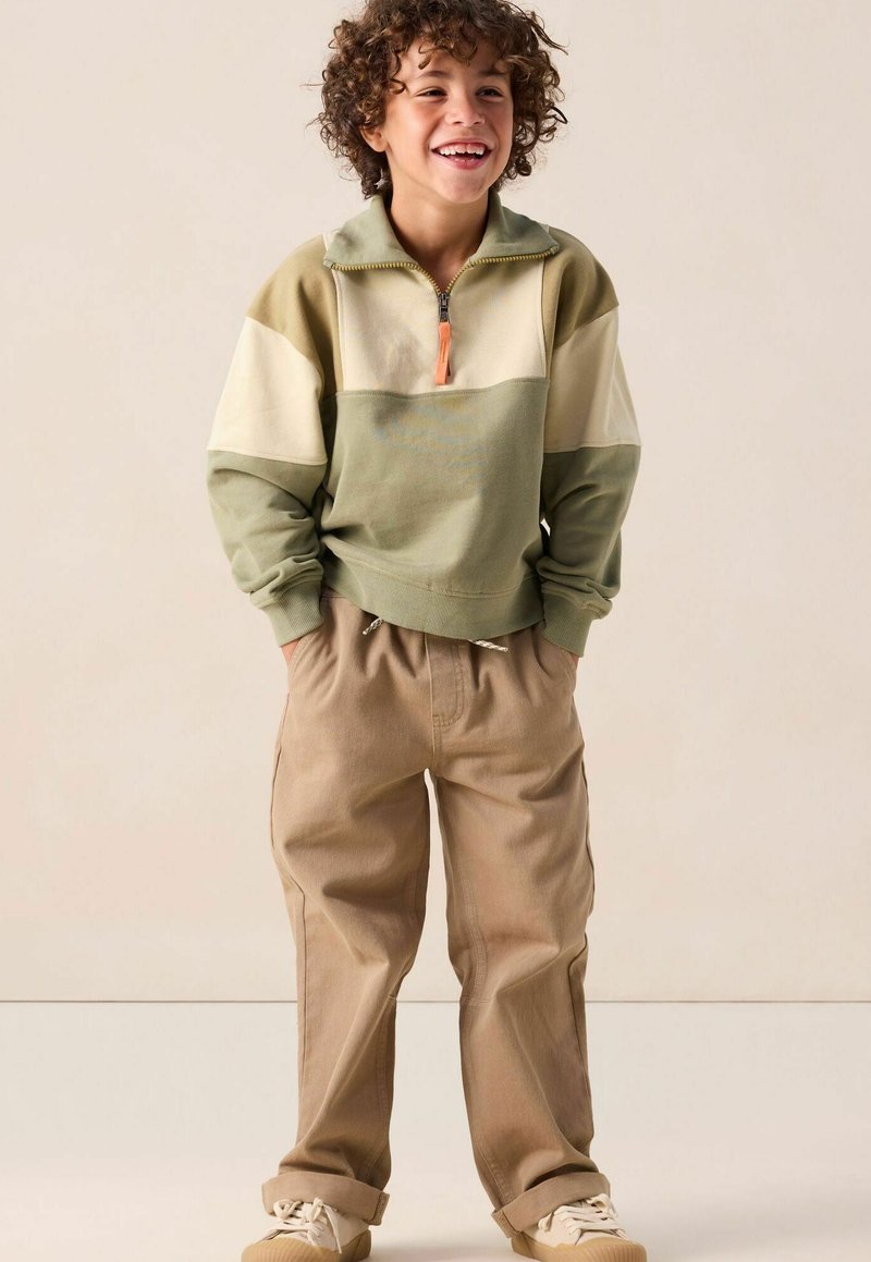 Groen en beige kleurblok sweatshirt met een ritskraag, gecombineerd met tan, relaxte broek. Zacht materiaal en details van een elastische tailleband.
