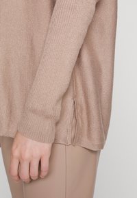 Nahaufnahme einer Hand neben einem beigen gestrickten Pullover mit seitlichem Schlitzdetail, kombiniert mit hellbraunen Hosen.