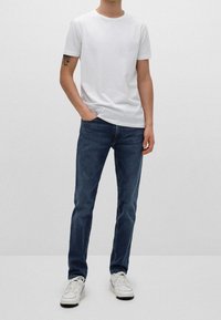 Muž v jednoduchém bílém tričku, tmavě modrých slim-fit džínech, bílých teniskách, s jednou rukou v kapse a viditelným tetováním na levém rameni.