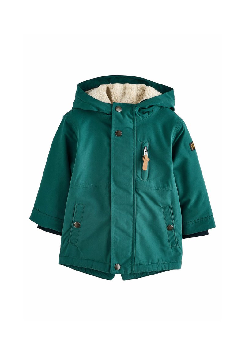 Veste verte isolée avec capuche, doublure en fausse fourrure, fermetures à boutons et à glissière, et grandes poches latérales. Elle présente un tissu texturé et des poignets contrastants en bleu marine.