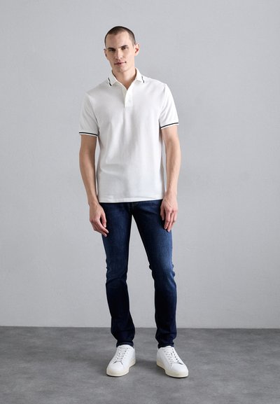 White polo shirt with black trim, paired with dark blue jeans. Model wearing white sneakers stands against a neutral gray background.