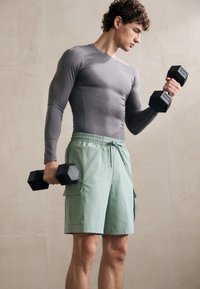 Pilka ilgomis rankovėmis sportinė marškinė ir šviesiai žali šortai su kišenėmis, laikantys juodus šešiakampius hantelius prieš neutralų foną.