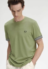 T-shirt in cotone verde con colletto a girocollo, logo ricamato nero e dettagli a righe bianco e nero sulle maniche corte. Tessuto morbido.