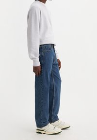 En person som står på sidan iförd en ljusgrå sweatshirt, blå raka jeans och vita sneakers med gröna sulor mot en vit bakgrund.