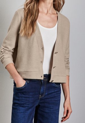 Frau trägt eine beige Strickjacke über einem weißen Oberteil und dunkelblaue Jeans, mit einer Hand in der Tasche, heller Hintergrund.