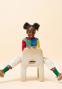 Sedia colorata in plastica riciclata con bordi arrotondati, caratterizzata da una texture a macchie. Bambino che indossa un maglione multicolore e scarpe con motivi.