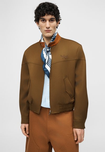Νέος άνδρας με σκούρα κατσαρά μαλλιά που φορά καφέ μπουφάν τύπου bomber, ανοιχτό μπλε πουκάμισο, μπλε και λευκή ριγωτή εσάρπα και καφέ παντελόνι που ταιριάζει.