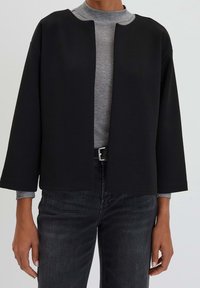 Svart kort jacka med en fyrkantig form, vida ärmar och öppen front. Bärs över en grå höghalshörning, i kombination med mörka jeans.